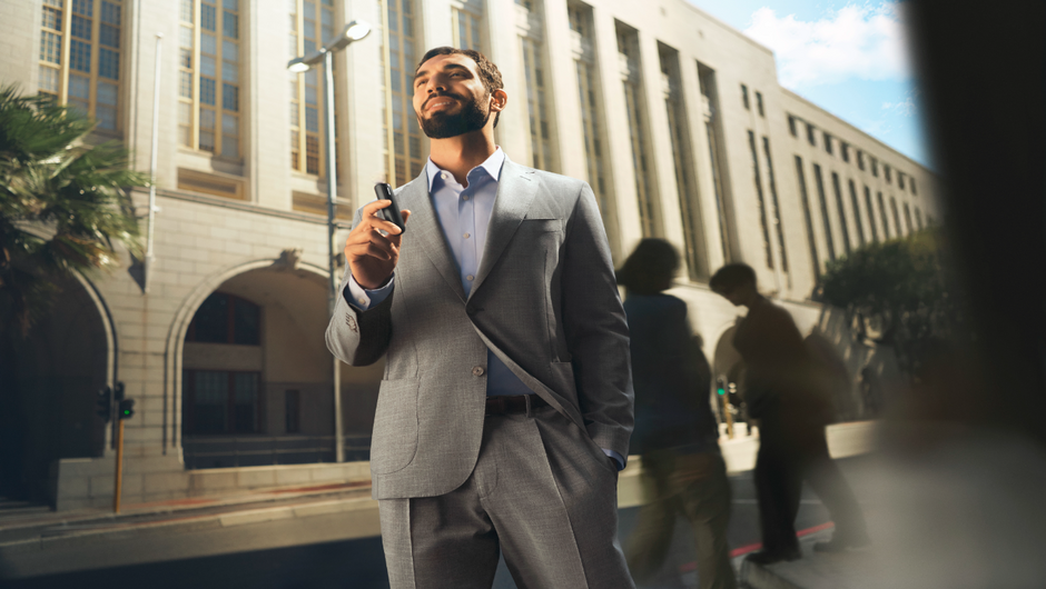 A confident man in a gray suit holds a Ploom Aura device, standing in front of a magnificent building.