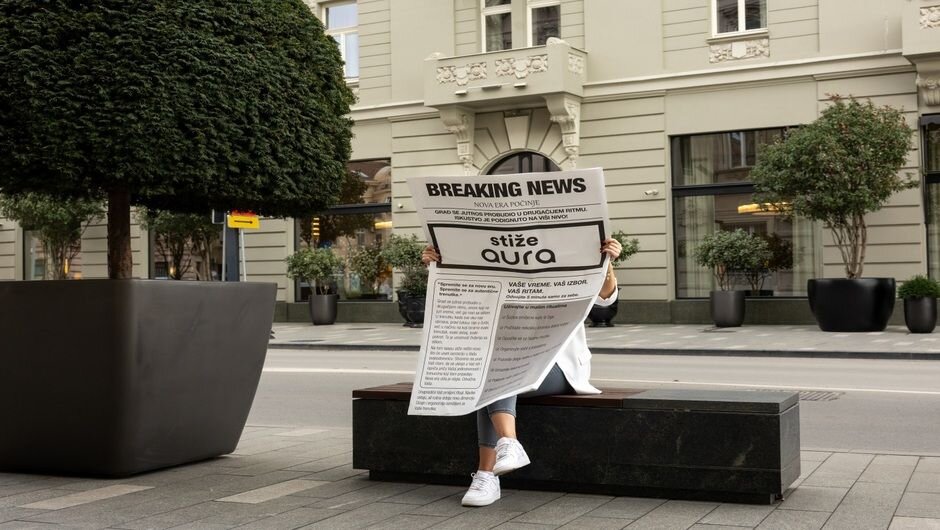 Person reads a large newspaper on a city bench, trees, road, historic building nearby.