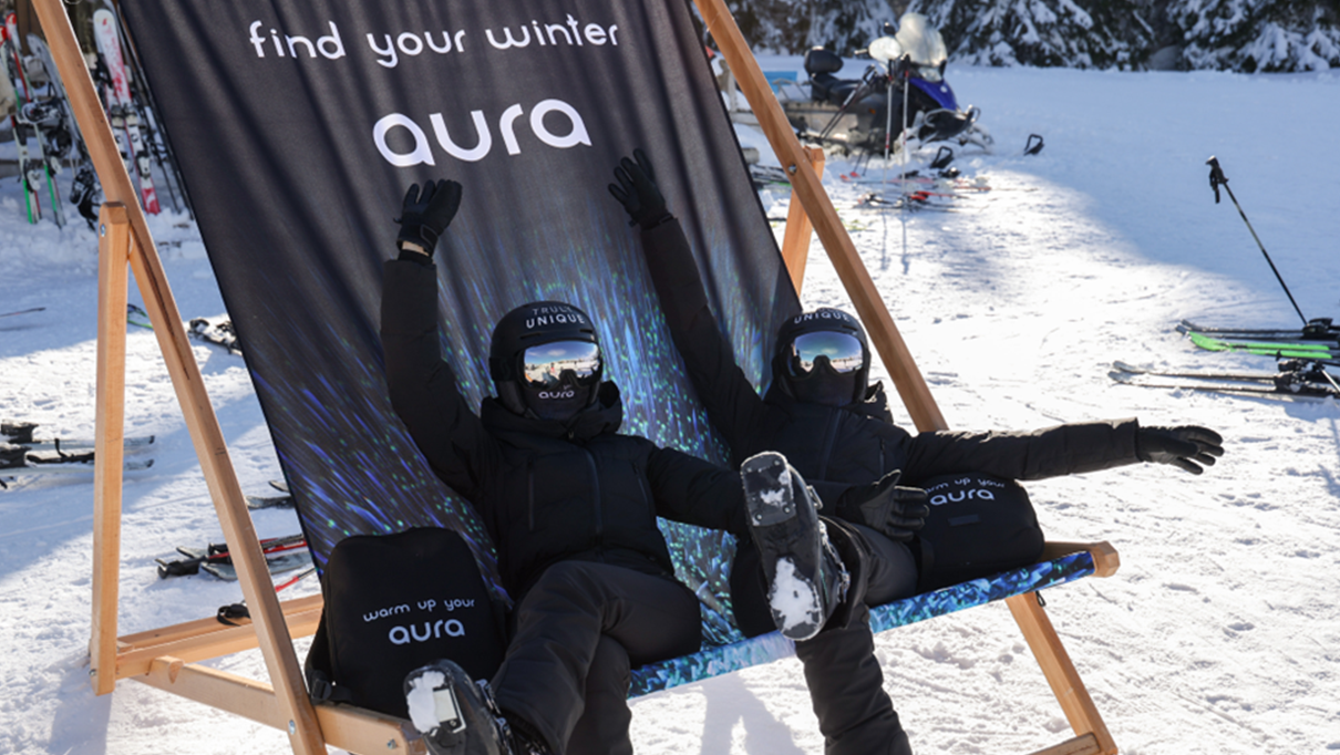 Two women in black ski outfits posing on a large AURA deck chair in the snow, in front of a branded “Find your winter aura” backdrop on a ski slope.