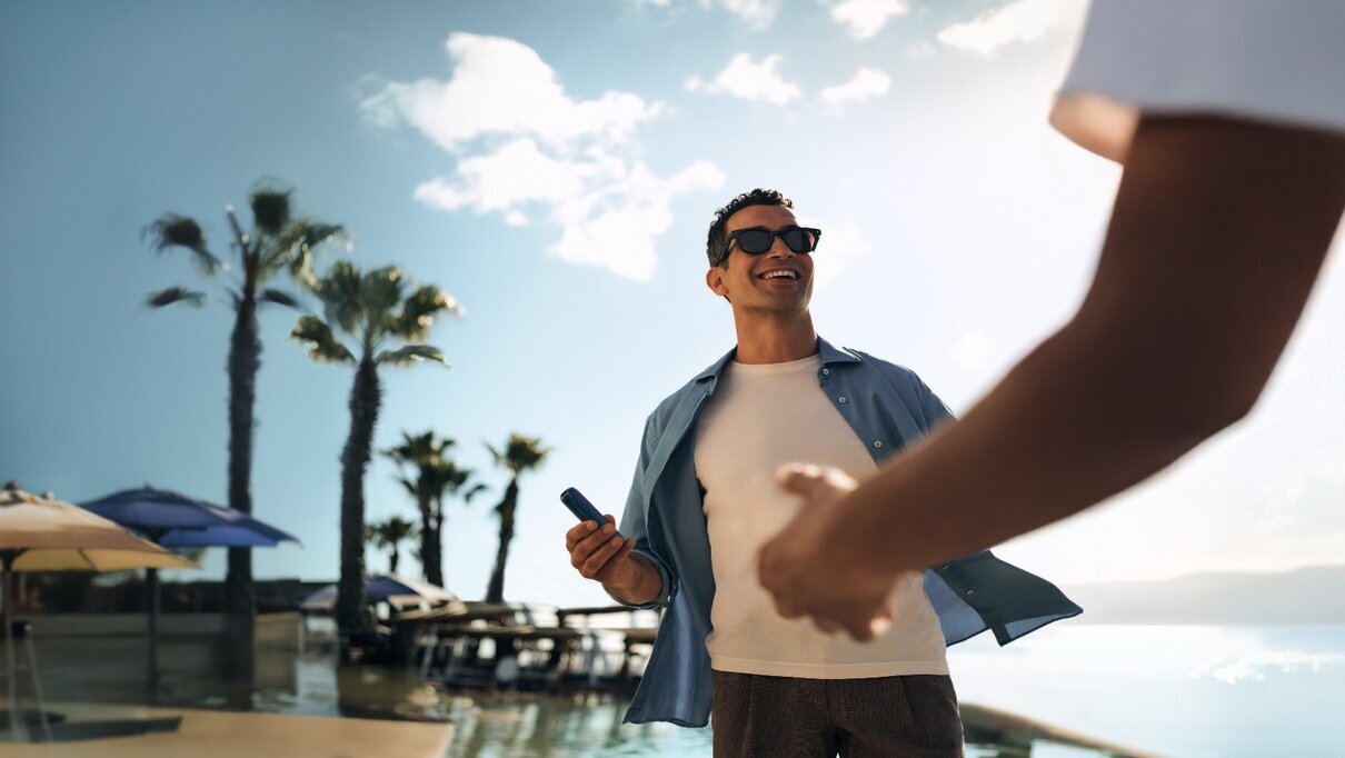 A man is smiling outdoors, holding a Ploom AURA device, with palm trees and a sunny sky around him.