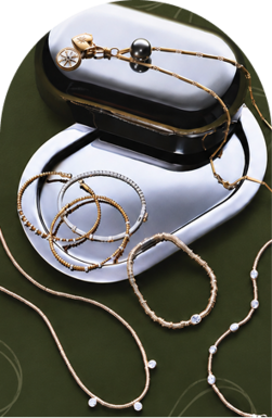 Gold and silver-toned necklaces and bracelets arranged around polished silver trays on patterned olive-green fabric.