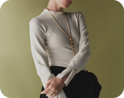 Model wearing a long gold pendant necklace, a delicate bracelet, and rings with a cream top and black trousers against an olive-green background.