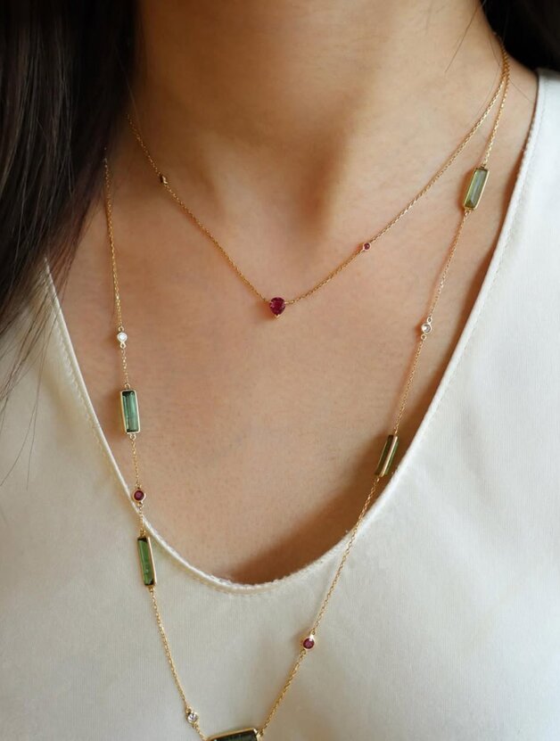 A person wearing a yellow gold ruby heart pendant necklace layered over a tourmaline and sapphire station necklace