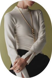 Model wearing a long gold pendant necklace, a delicate bracelet, and rings with a cream top and black trousers against an olive-green background.