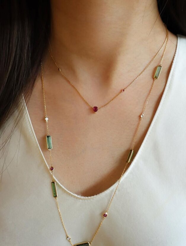 A person wearing two layers of dainty yellow gold necklaces featuring multicolored gemstones