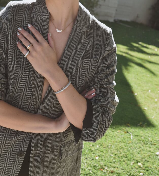 A woman wearing a dark grey herringbone wool blazer with a set of yellow gold, diamond-embellished necklace, rings, ear cuff and bangle