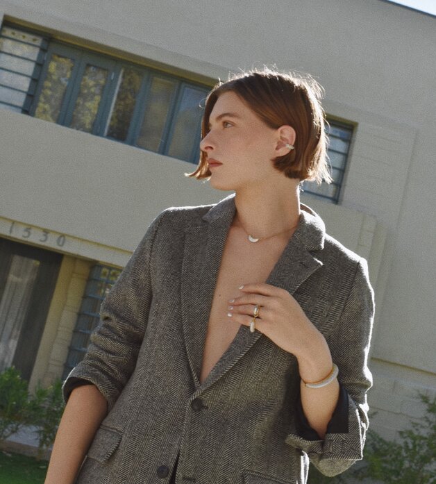A woman wearing a dark grey herringbone wool blazer with a set of yellow gold, diamond-embellished necklace, rings, ear cuff and bangle