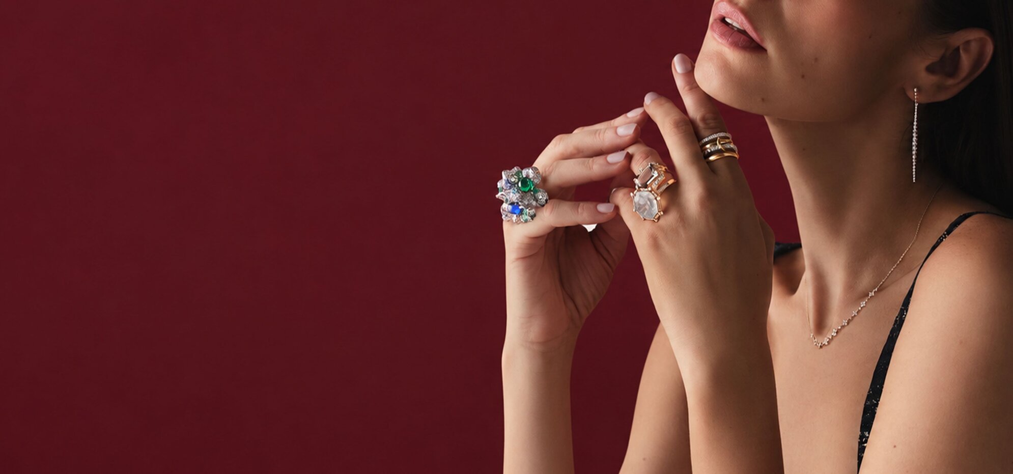 Model wearing statement gemstone rings, a delicate necklace, and drop earrings against a deep burgundy background.