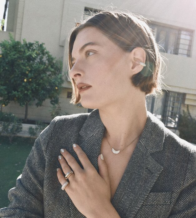 A woman wearing a dark grey herringbone wool blazer with a set of yellow gold, diamond-embellished necklace, rings, ear cuff and bangle