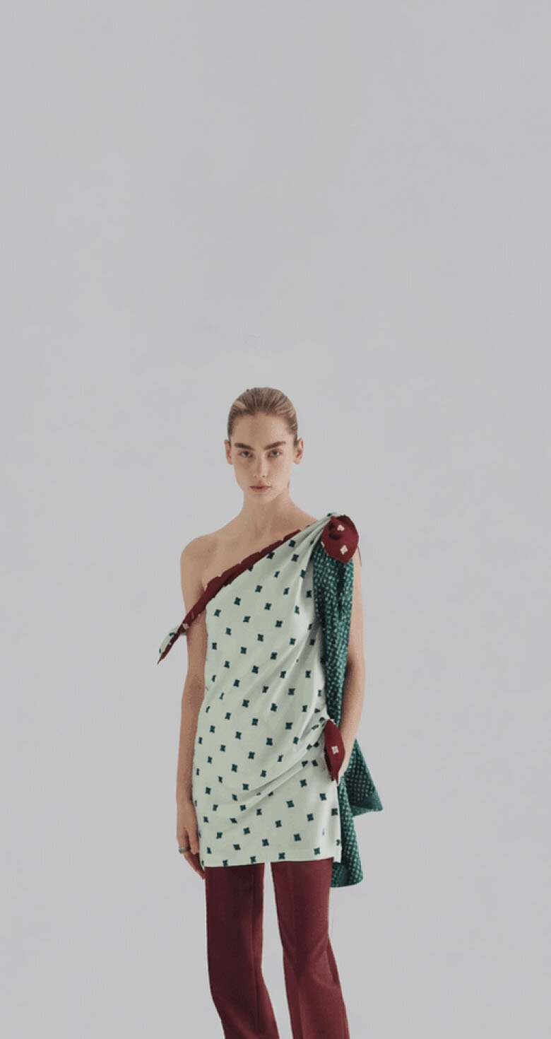 An image of a woman wearing a light green asymmetric one-shoulder printed mini dress over maroon trousers