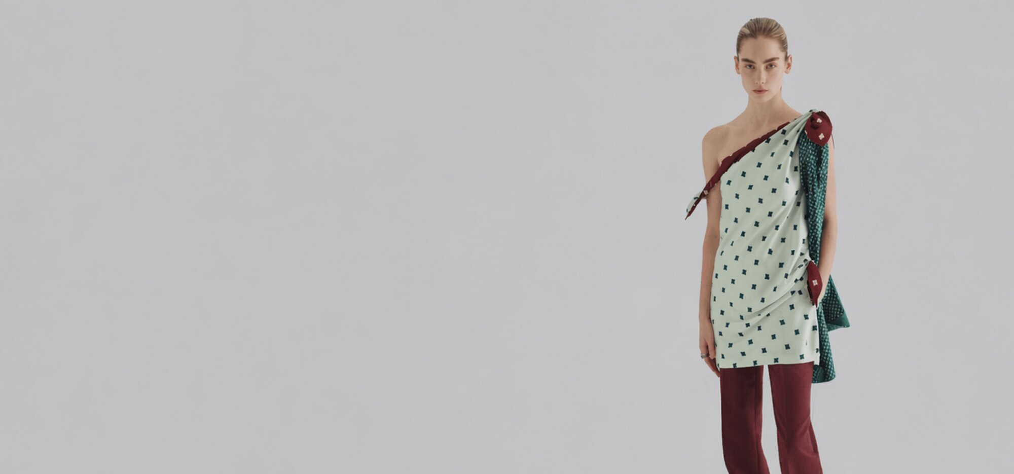An image of a woman wearing a light green asymmetric one-shoulder printed mini dress over maroon trousers