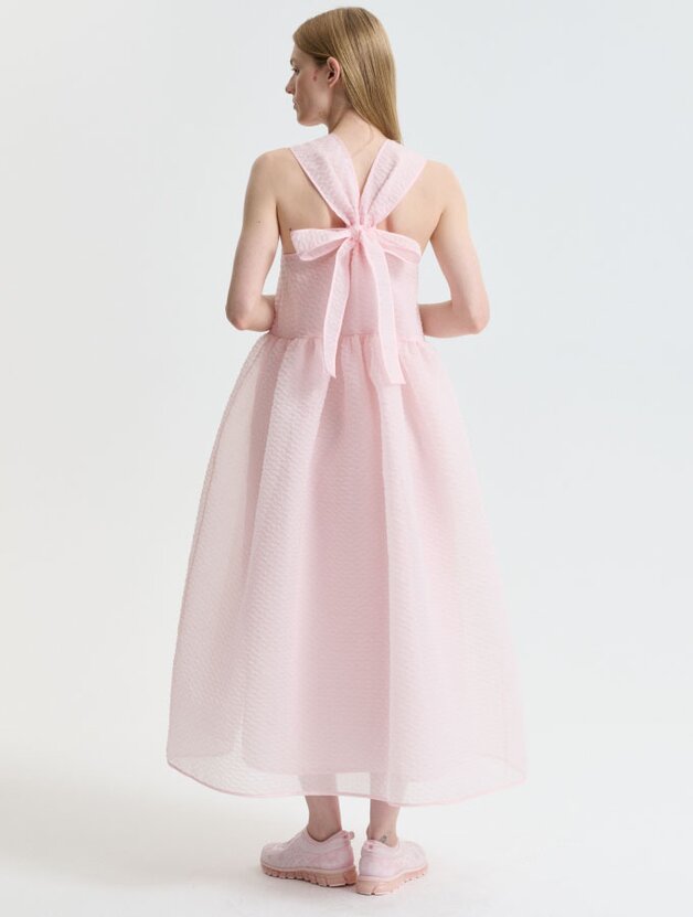 A woman wearing a light pink sleeveless matelassé midi dress featuring a bow detail at the back