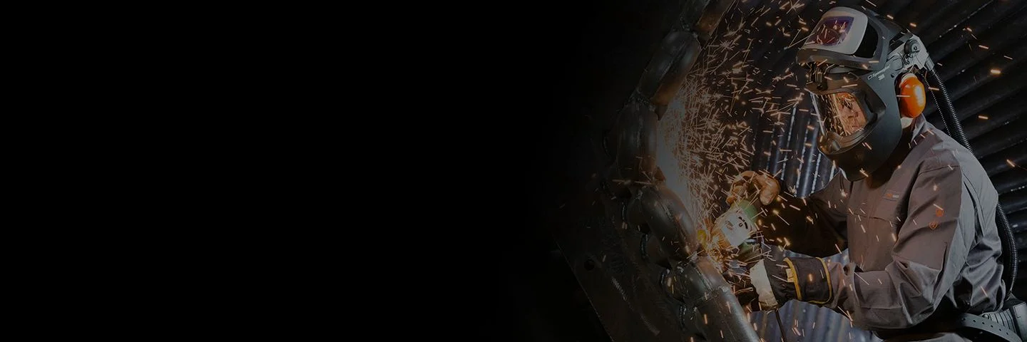 A man wearing 3M protective face and bodywear grinds metal, sparks flying.