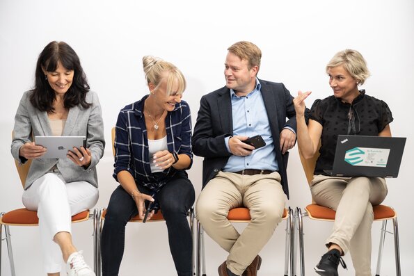 Drei Frauen und ein Mann sitzen in einer Reihe auf Stühlen mit orangen Sitzpolster und haben eine gute Zeit. Sie lachen und halten Smartphones, Tablets und Laptops in der Hand. 