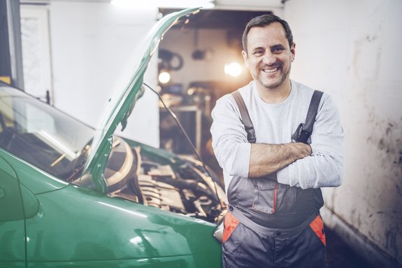 Im Vordergrund steht ein Mann mittleren Alters, der in die Kamera lächelt. Er trägt eine dunkelgraue Latzhose als Arbeitskleidung und befindet sich in einer Werkstatt. Er lehnt sich an ein grünes Auto, welches die Motorhaube offen hat.