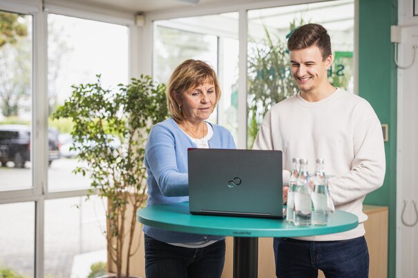 Eine ältere Frau und ein junger Mann stehen an einem Hochtisch und schauen auf ein Laptop.