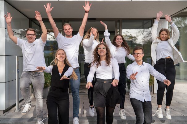 Gruppenfoto von Sellwerk Mitarbeitern die beim Standort Nürnberg vor dem Eingang des Bürogebäudes in die Luft springen und die Hände heben. 