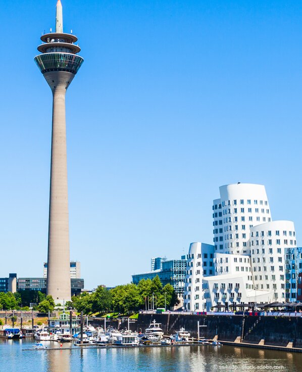 Rheinturm in Düsseldorf, daneben ein weißes Gebäude mit vielen Fenstern und vielen Booten im Vordergrund.