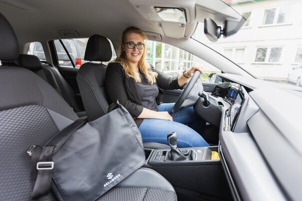 Ein Frau sitzt im Auto und lächelt in die Kamera. Auf dem Beifahrersitz ist eine schwarze Tasche mit dem Sellwerk Logo.