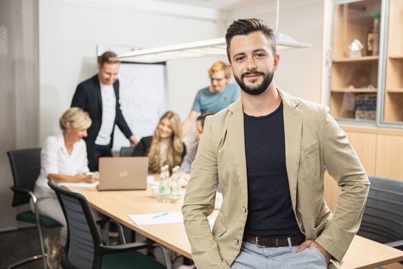 Ein SELLWERK Mitarbeiter einem Anzug, lächelt in die Kamera, während im Hintergrund andere SELLWERK Mitarbeiter etwas besprechen.