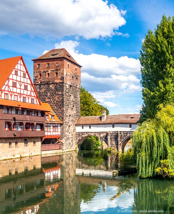 Alte Brücke über dem Fluss Pegnitz in Nürnberg daneben ein Fachwerkhaus und verschiedene Bäume.