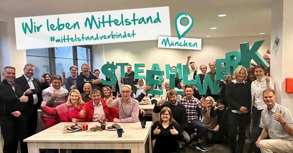 Im Vordergrund steht ein Mann mittleren Alters mit weißen Hemd und grauen Blazer, der in die Kamera lächelt und die Arme verschränkt hat. Im Hintergrund sind unscharf das Sellwerk Logo, sowie ein Mann und eine Frau zu sehen.