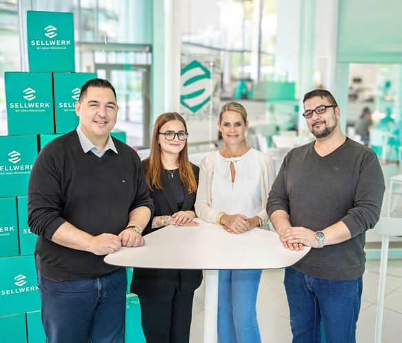 Vier Personen stehen an einem Stehtisch und grinsen in die Kamera.