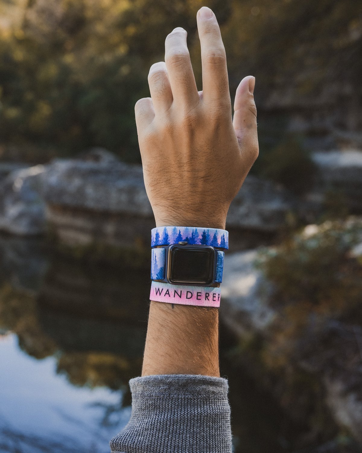 An arm raised with a ZOX wristband and ZOX watchband in front of a mountain creek.