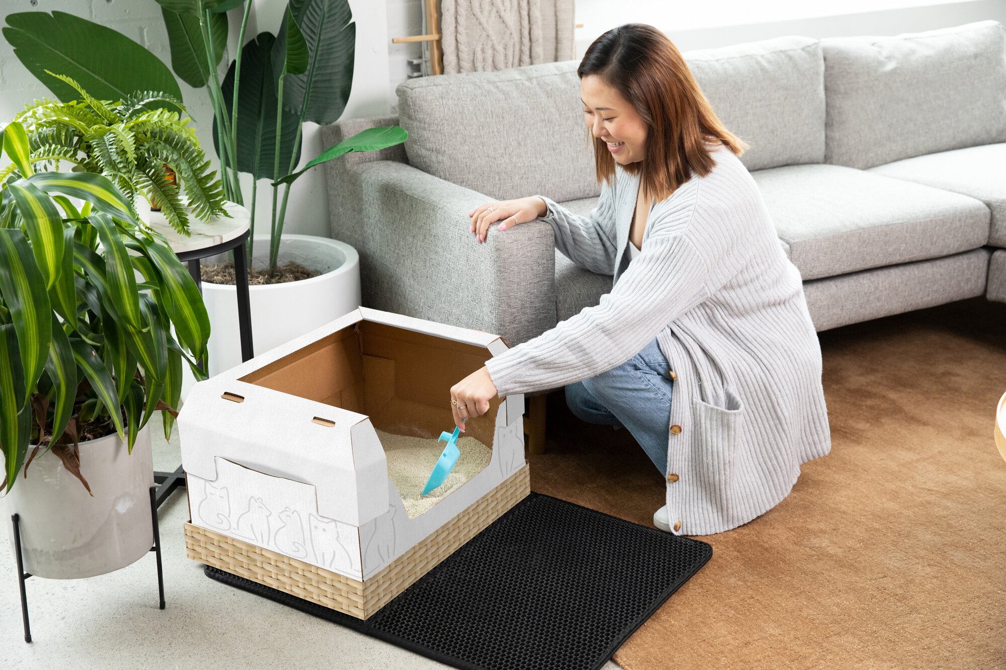 Woman sitting on a gray couch with her black and white cat. Kitty Poo Club disposable litter box is placed next to the couch.