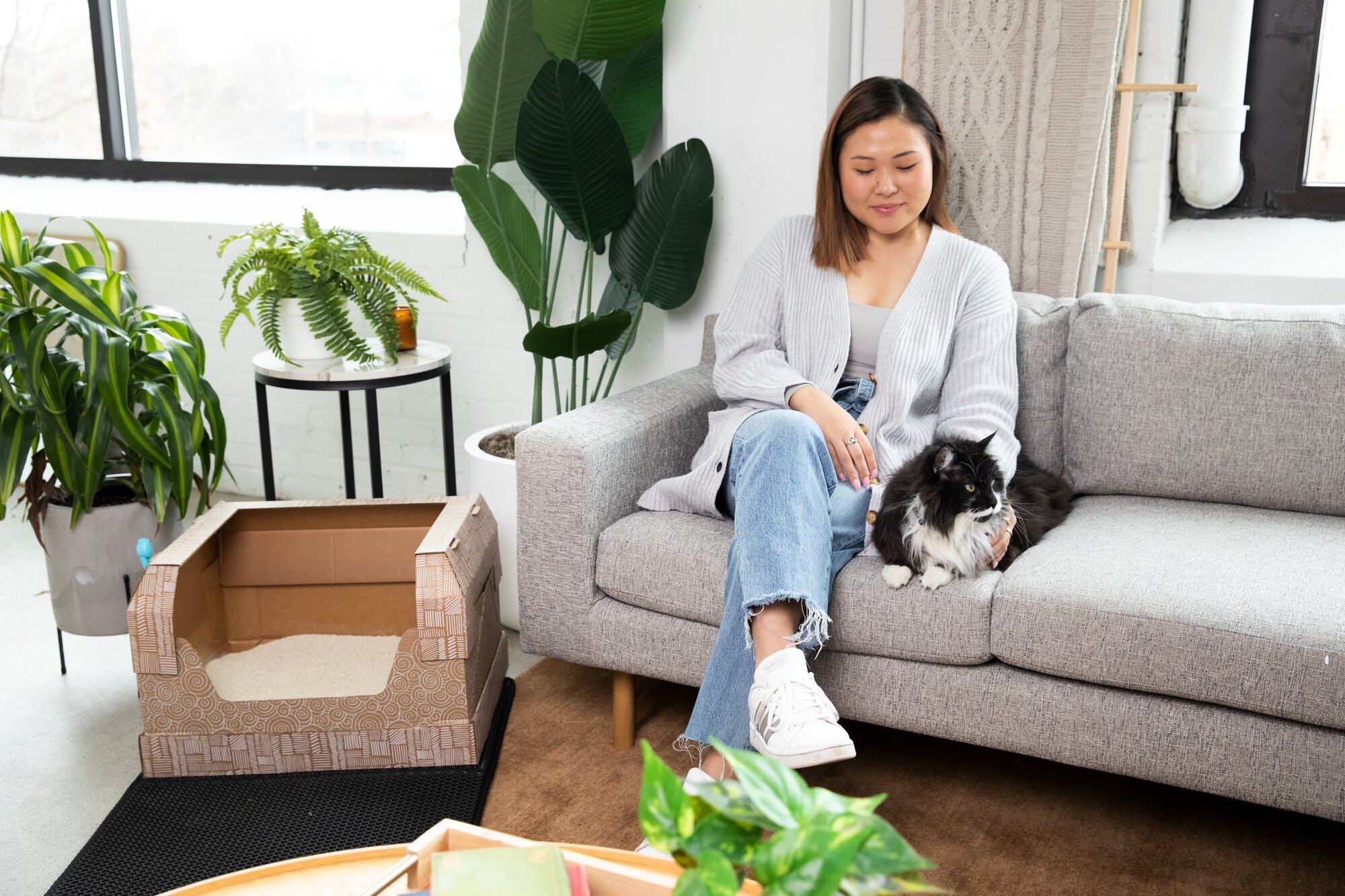 Woman sitting on a gray couch with her black and white cat. Kitty Poo Club disposable litter box is placed next to the couch.