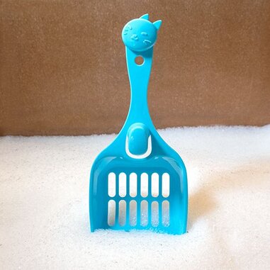 A bright blue cat litter scoop with a cute cat face on the handle, standing upright in a bed of white cat litter against a neutral brown background.