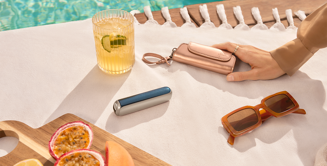 Accessori Ploom e dispositivo Ploom Aura appoggiati su un telo a bordo piscina, con drink e occhiali da sole.