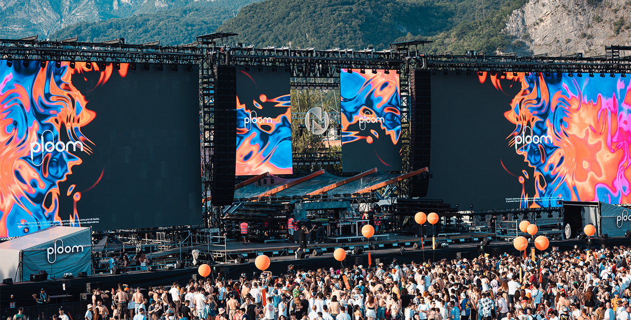 Grande palco brandizzato Ploom durante un festival all’aperto, con folla, schermi scenografici e montagne sullo sfondo.