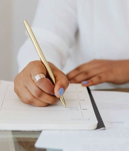 Close-up of a woman in a white long-sleeve shirt writing in a daily planner with a pen, wearing a ring and with painted nails.