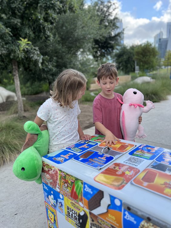 Two young children visit an ice cream stand with our Toby the Turtle and Lola the Axolotl plushes