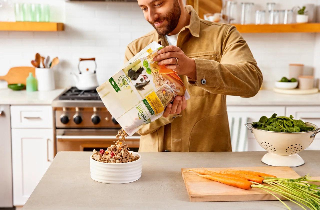 Man in a kitchen pours Freshpet dog food from a bag into a bowl on the counter.