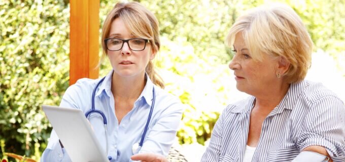 Elderly woman sitting in the garden with home health care nurse and consulting. Middle age caregiver holding in hand digital tablet and giving advice to her patient.