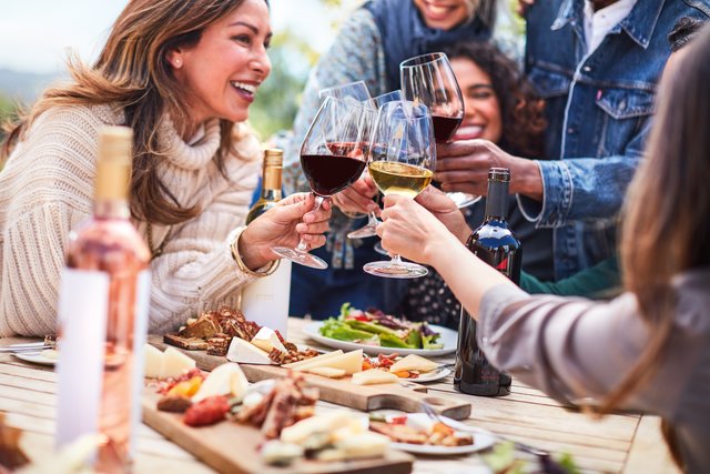 A group of smiling friends toast glasses of red and wine over an outdoor table of vibrant, fresh food.