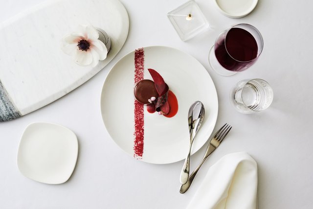  A glass of red wine and a plate with a glossy chocolate gâteau with deep red garnishes.