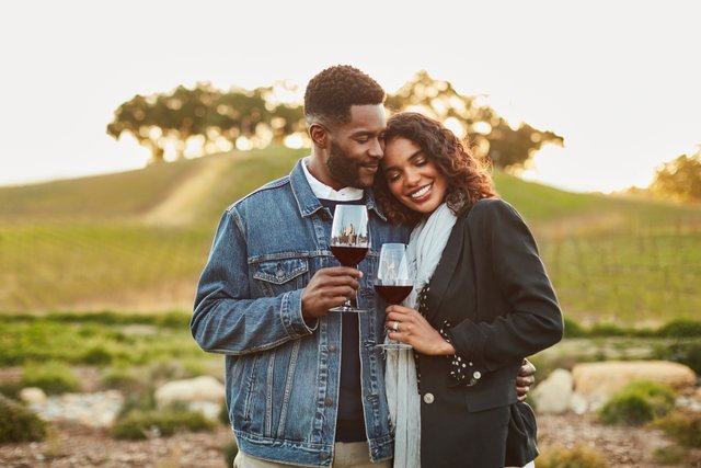 A smiling woman leans her head on a man's shoulder, each holding a glass of red wine, sunlit trees in the background.