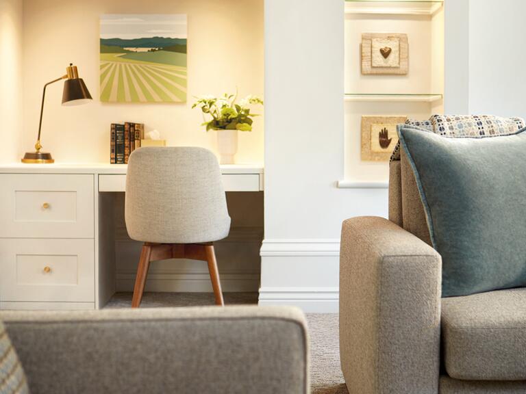 A room with white walls highlighting a small white desk, a shelf, and two beige furniture pieces with a slate grey pillow