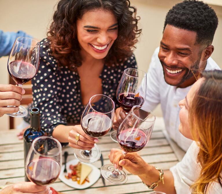 Three, grinning friends toast glasses of red wine over a wooden table with fresh food.