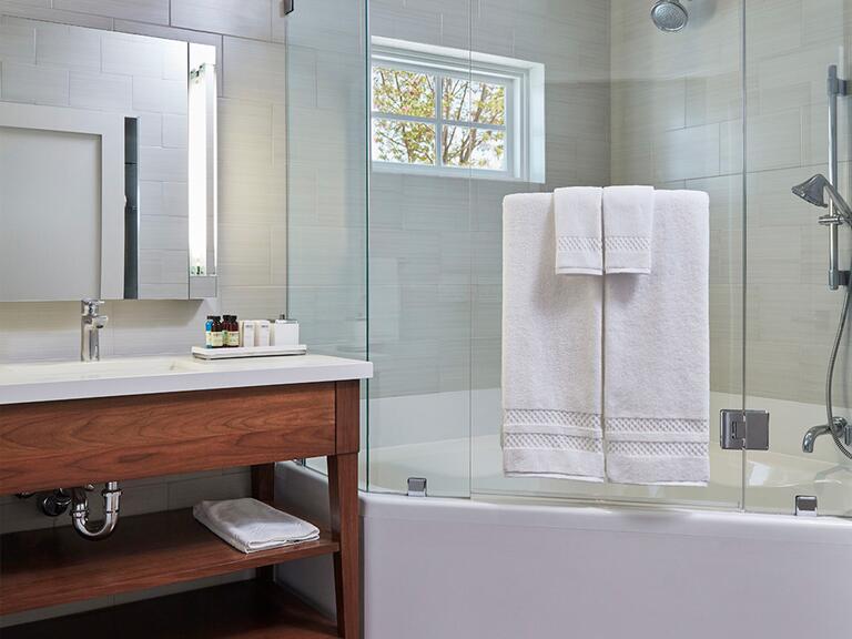 A bathroom with beige tile walls, a mirror hanging above a white countertop and wood sink area, and a full shower on the left with a window.