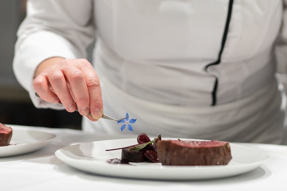 A small, blue flower is placed with tweezers on a steak dish for garnish.