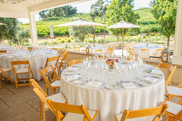 Four, large, round tables set for 12 sit in the patio shade, overlooking the vineyard. 
