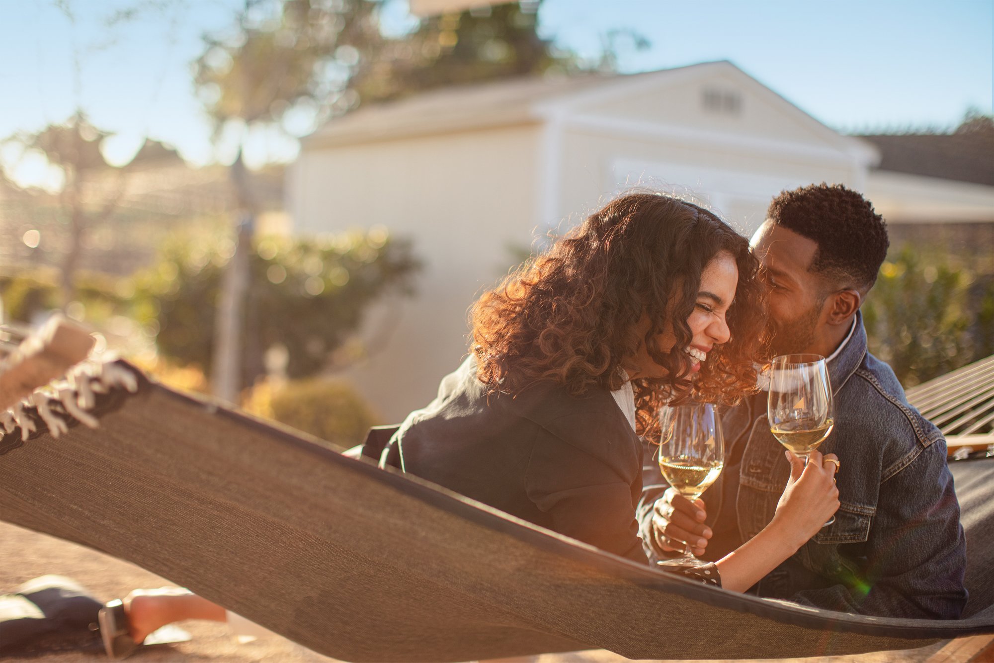 A woman, holding a glass of white wine and laughing, sits in a hammock with a man who whispers in her ear.