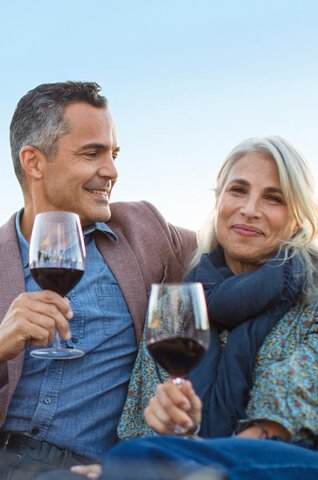 A woman and man are leaning into each other laughing, holding red glasses of wine and laughing