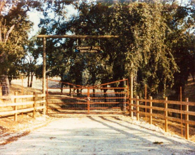 An old image of a ranch-like gate with a sign that reads "Chimney Rock Vineyards" and large trees all around it.