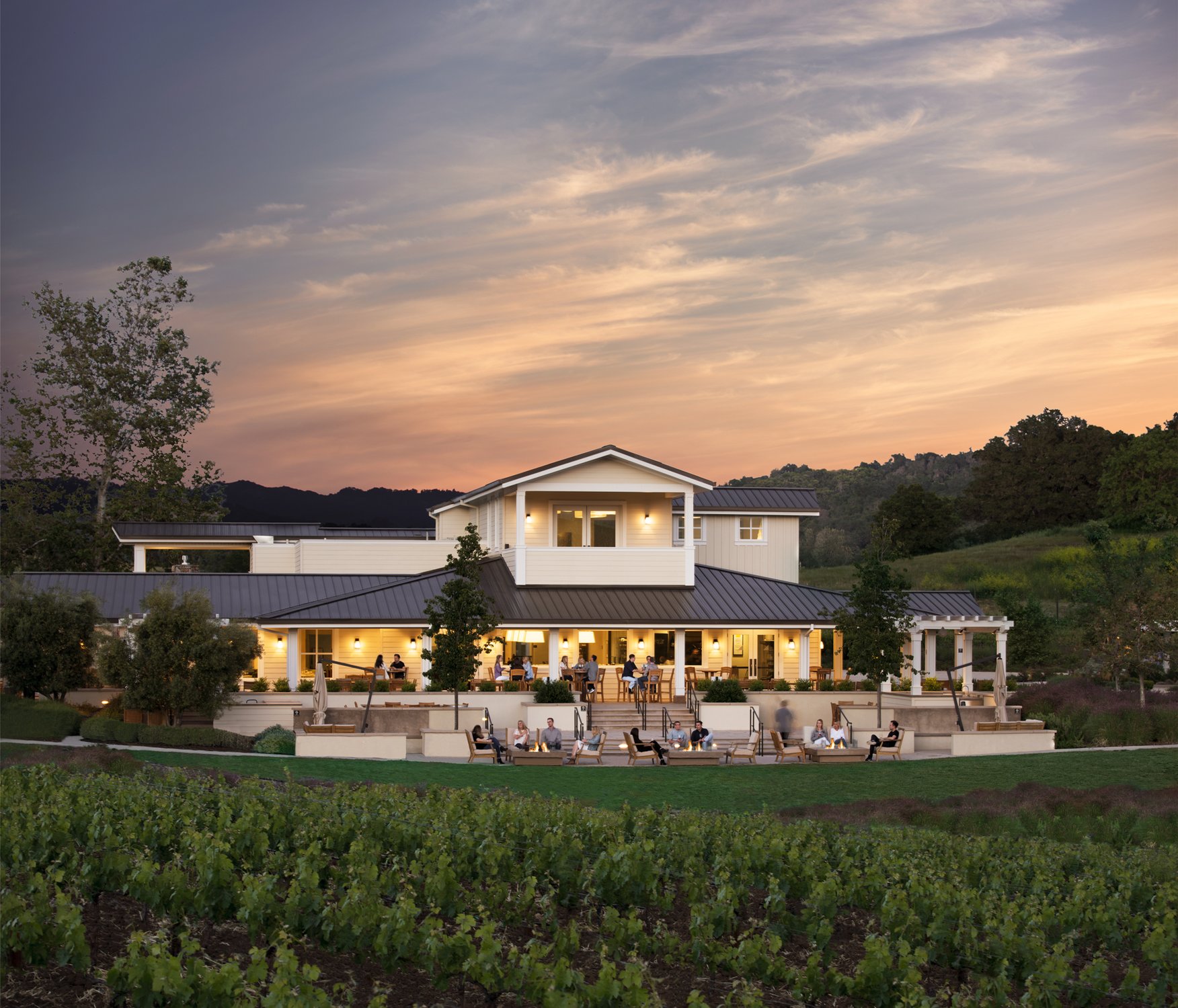 Guests lounge outside the warmly-lit white buildings of the JUST Inn sit under a peach-colored, dusk sky.