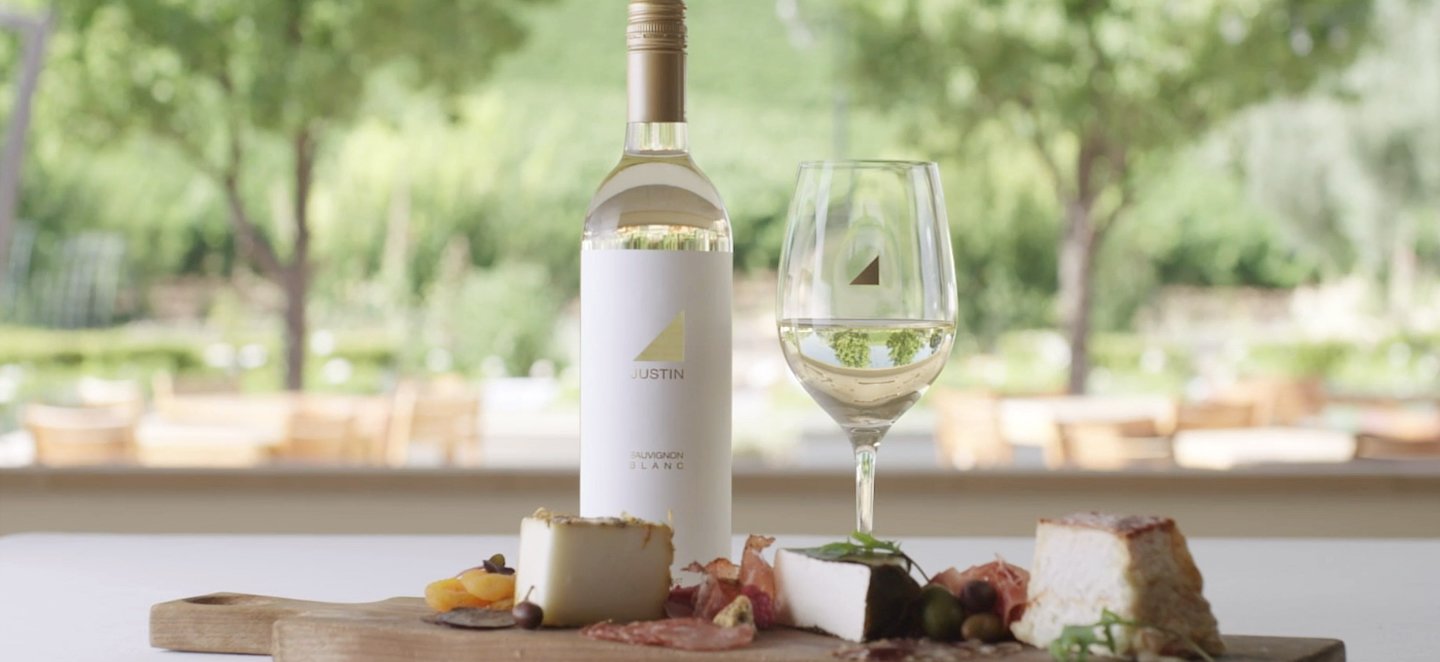 A bottle and glass of JUSTIN Sauvignon Blanc with assorted charcuterie, the patio and sage green landscape behind them. 